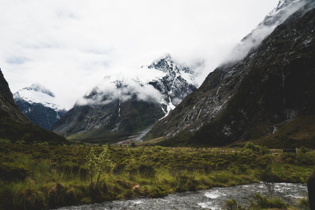Snow-capped mountain peaks above green valley with swirling clouds and flowing stream landscape Wallpaper