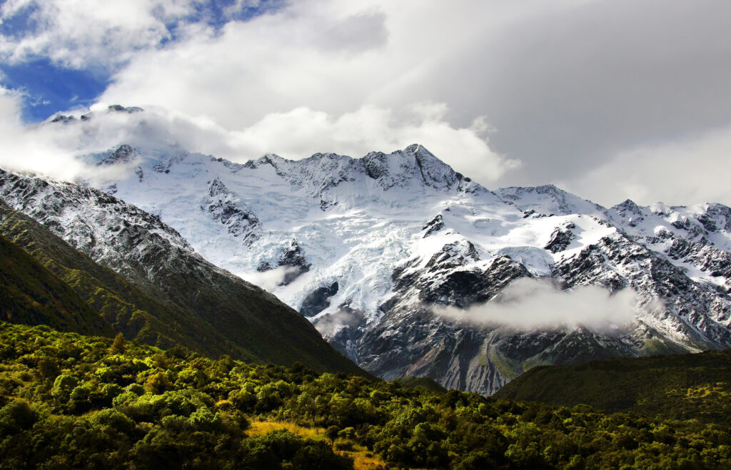 Snow capped mountains with dramatic clouds lush green trees and rolling hills wallpaper background