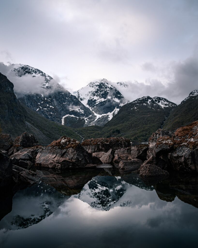 Snow-capped mountains reflected in a serene lake with rugged rocks and dramatic clouds wallpaper