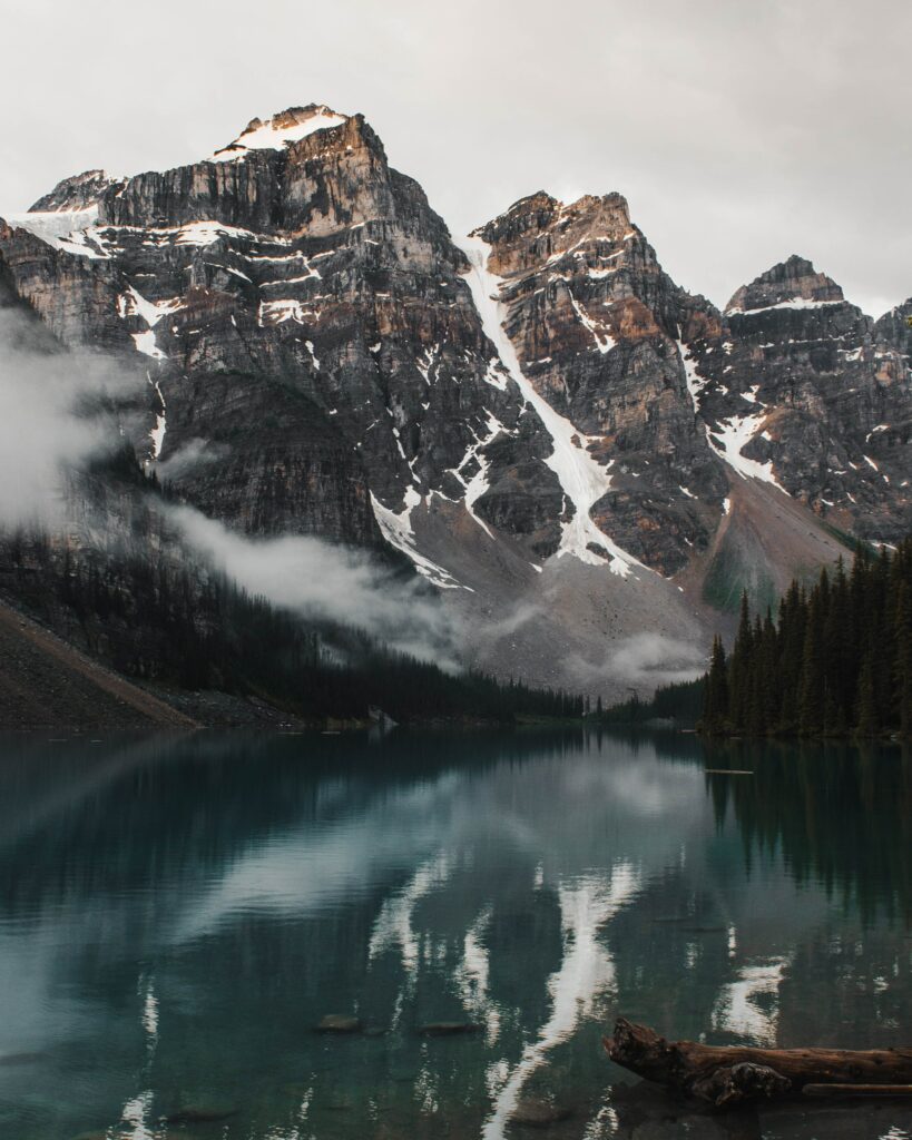 Snow-capped mountains reflected in calm lake surrounded by evergreen trees and misty peaks Wallpaper