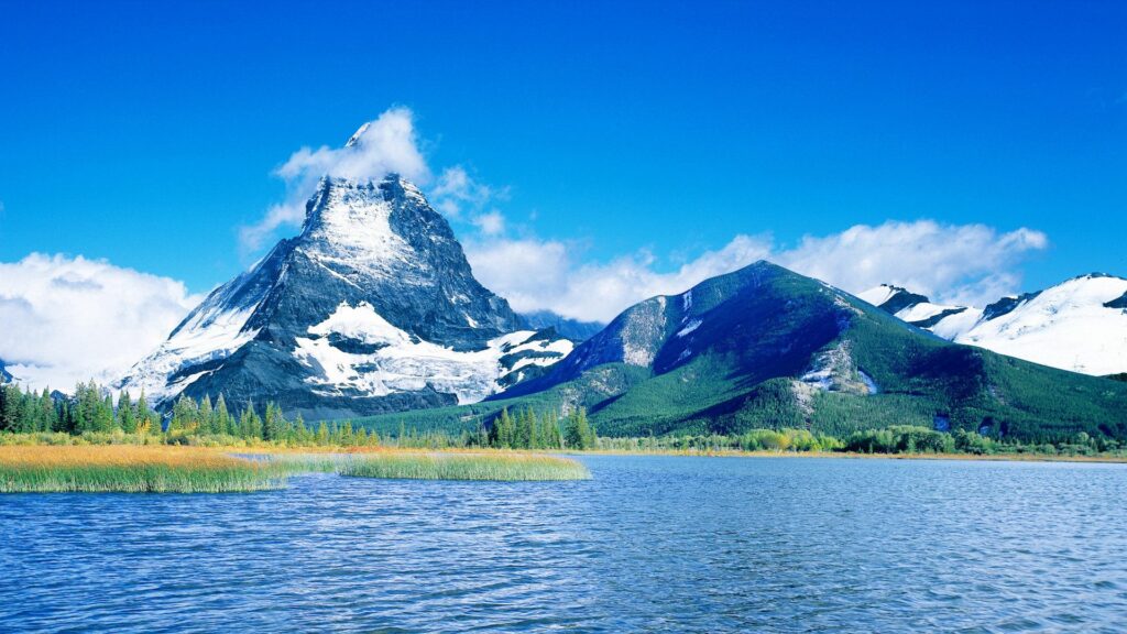 Snow-capped mountains under blue sky reflected in calm lake with lush green trees shore wallpaper