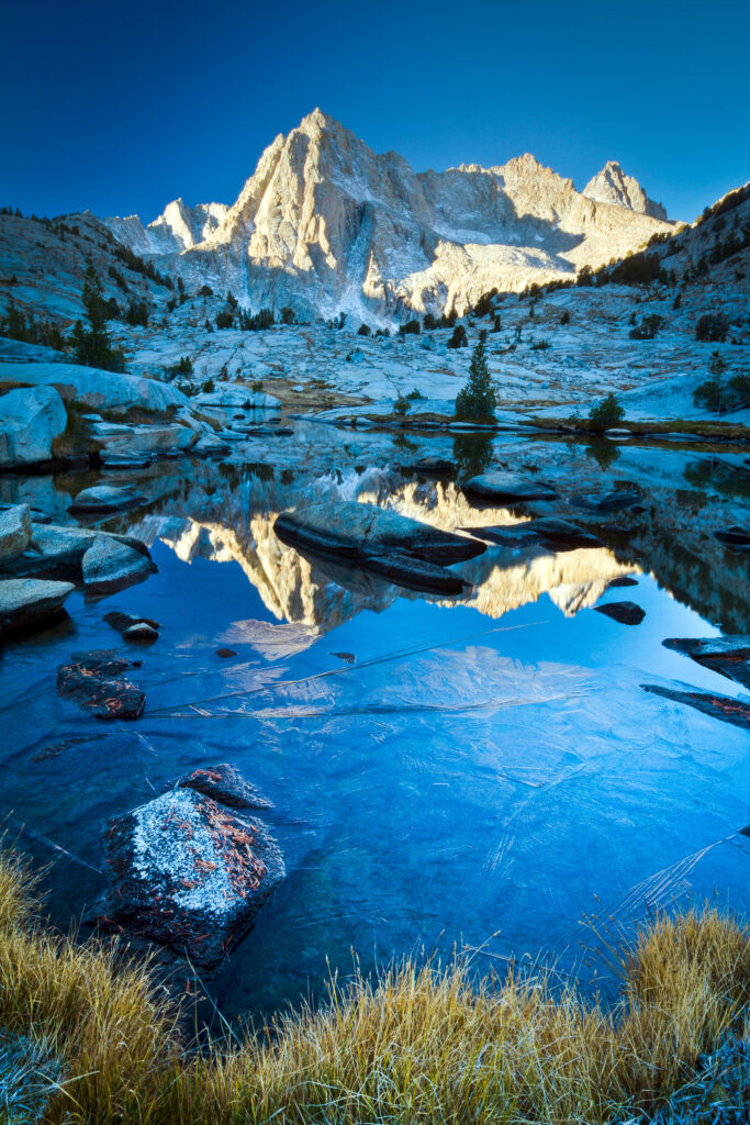 Snow-capped mountains reflected in clear water with vibrant blue sky and rocky grassy terrain Wallpaper