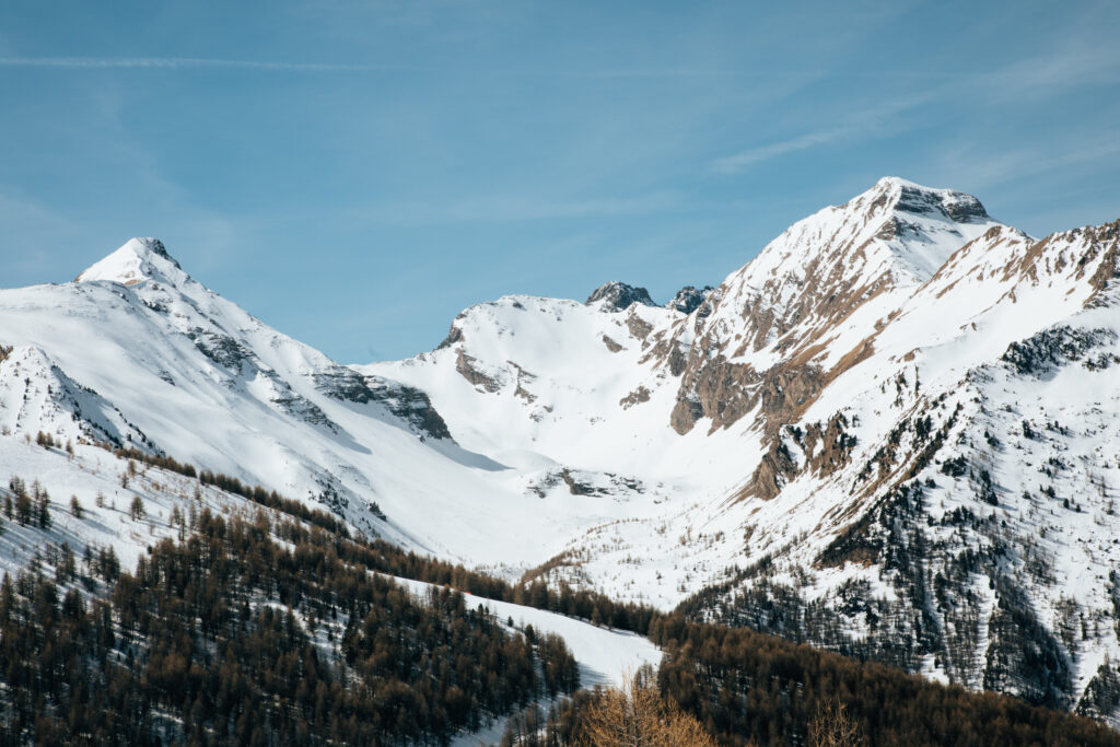 Snow-covered alpine mountains with rugged slopes and scattered trees under a clear blue sky Wallpaper