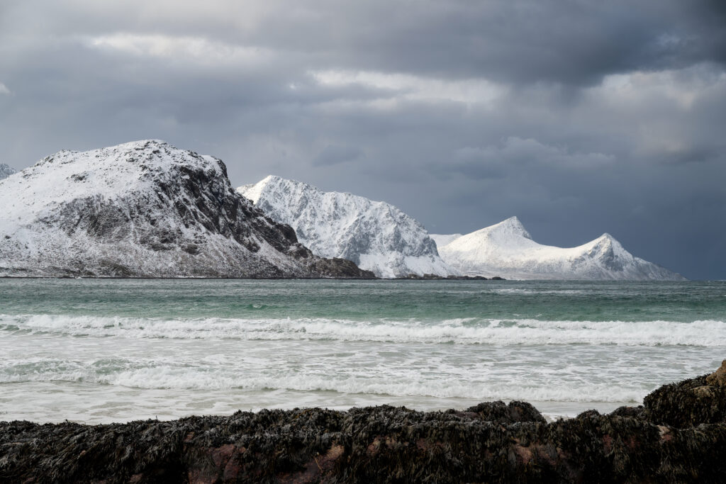 Snow-covered mountains meet turquoise sea under brooding grey clouds dramatic coastal landscape Wallpaper