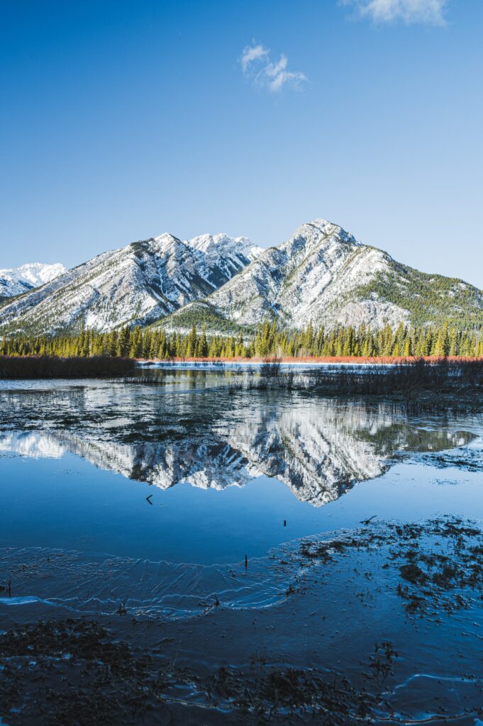Snow-dusted mountains reflected in a clear lake with evergreens under a bright blue sky wallpaper