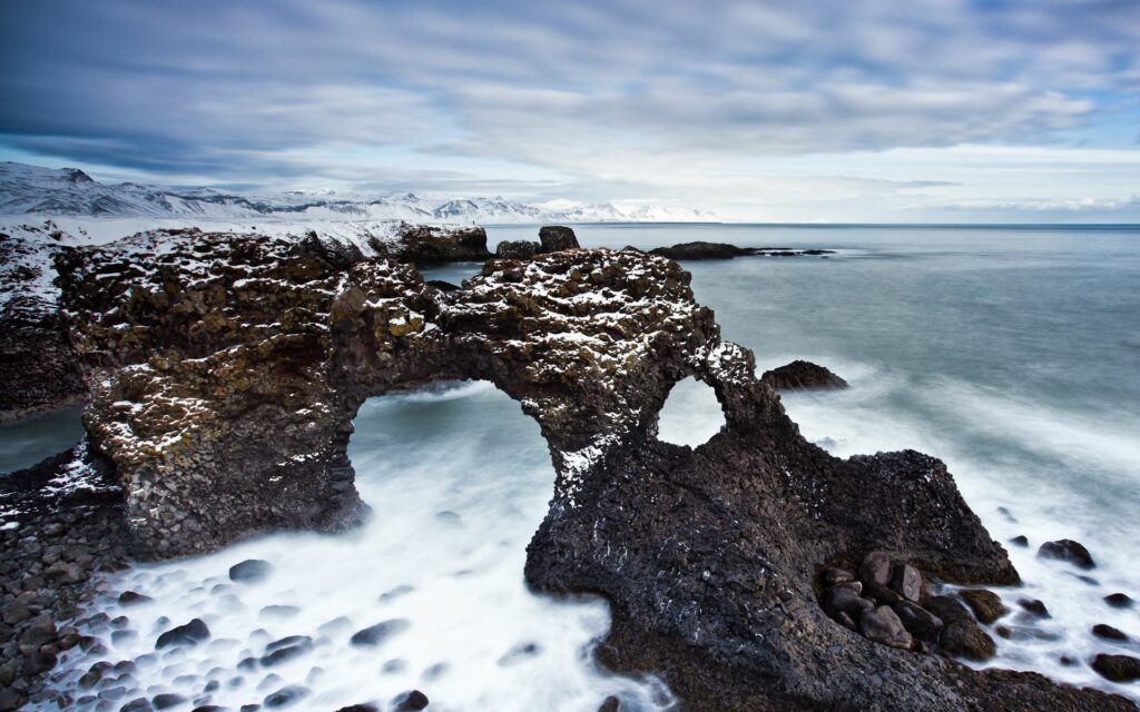 Rugged stone arches on snowy coastline with misty waves and cloudy sky wallpaper background