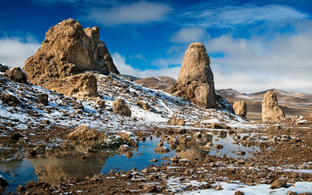 Snow-dusted stone formations under blue sky with reflective puddle in barren landscape wallpaper