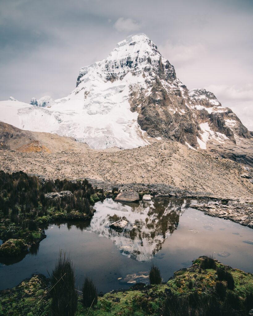 Snow-capped mountain peak reflected in tranquil lake surrounded by lush greenery and rocks Wallpaper