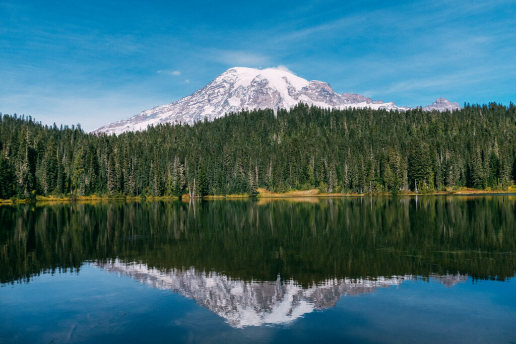 Snow-capped mountain above evergreen forest reflected in clear lake under bright blue sky wallpaper