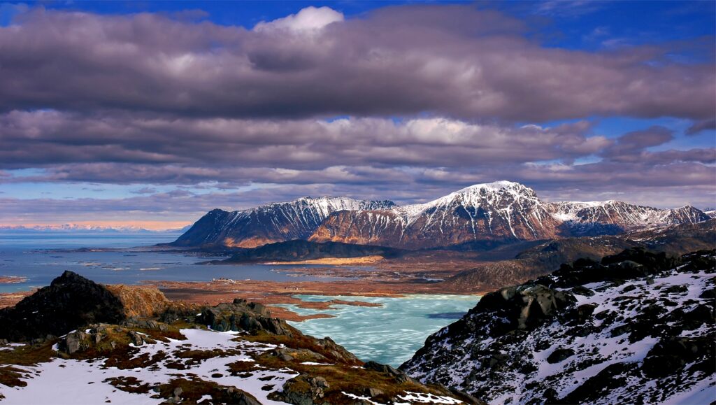 Snow-capped rugged mountains under dramatic swirling clouds with icy waters and blue sky wallpaper