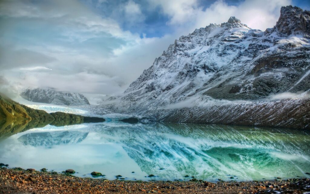 Snow-capped mountains reflect in crystal-clear lake with floating ice rugged stones and clouds Wallpaper