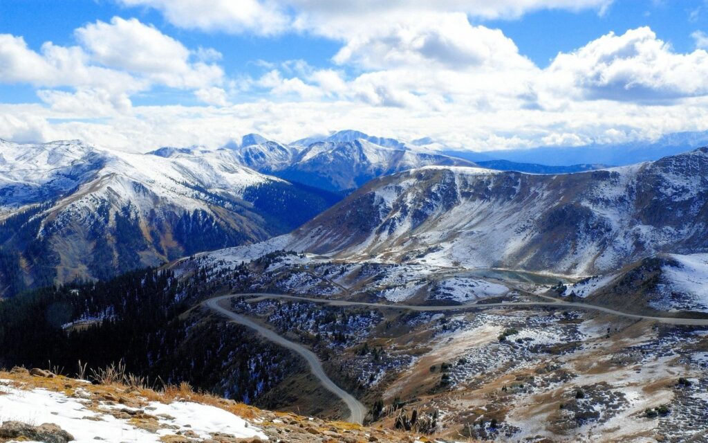 Snow-capped mountains with winding road under bright blue sky high-altitude rugged landscape wallpaper