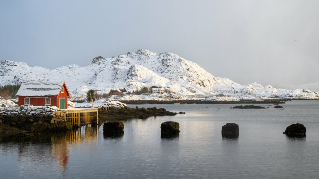 Serene winter landscape with snow-capped mountains reflected in calm lake and red cabin Wallpaper