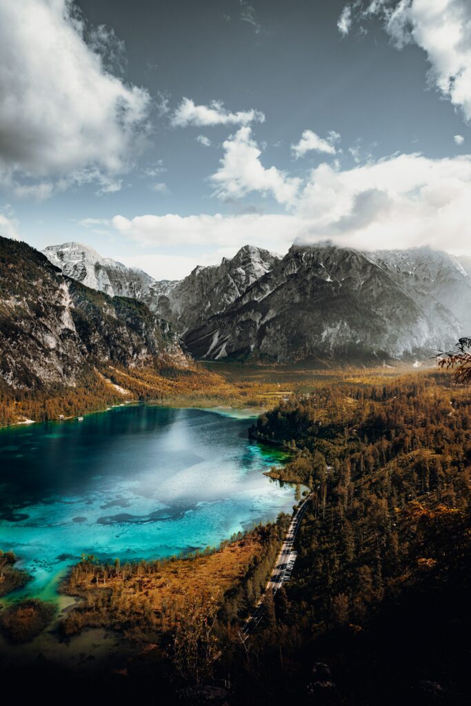 Snow-capped mountains reflected in turquoise lake with autumn forest and winding road landscape Wallpaper