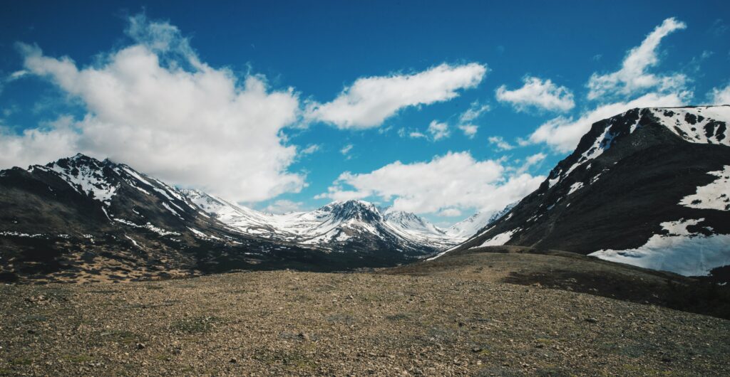 Snow-capped rugged mountains under vibrant blue sky with fluffy clouds and earthy foreground wallpaper
