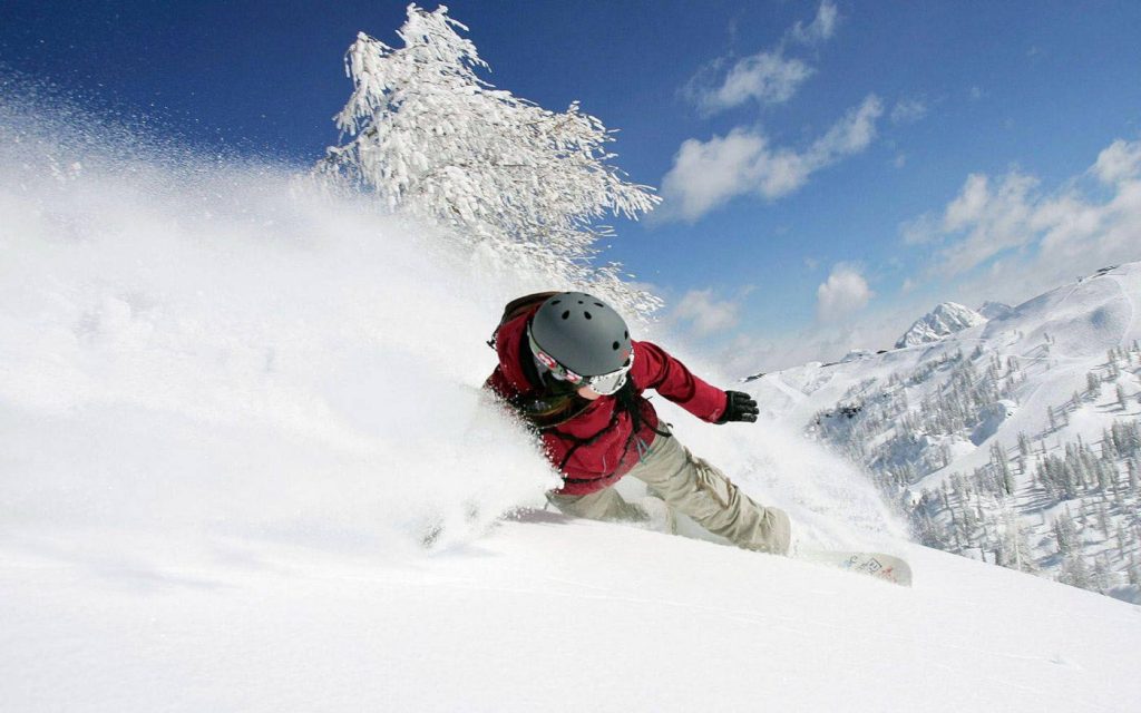 Snowboarder in red jacket carving fresh snow on sunny mountain slope with frosted trees background Wallpaper