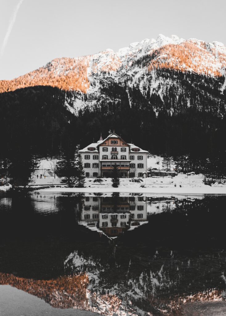 Charming snowy house reflected in tranquil lake with sunlit snowy mountains landscape wallpaper