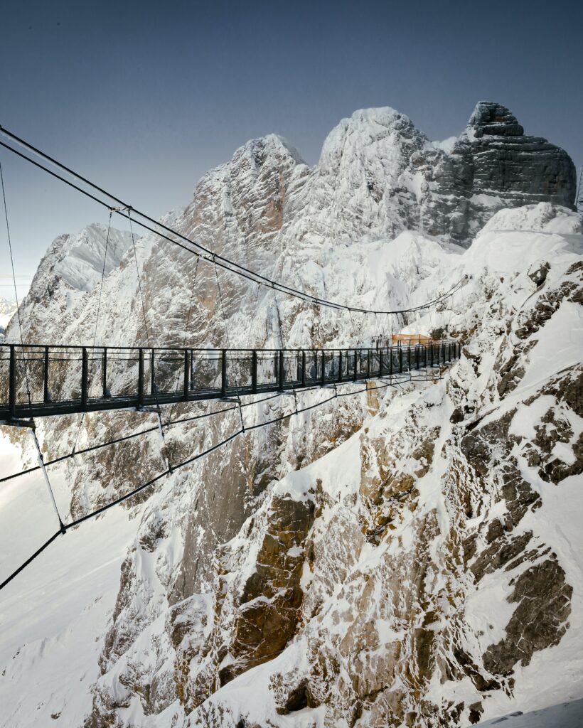 Winter wallpaper of suspension bridge over snow-covered rugged mountains under blue sky