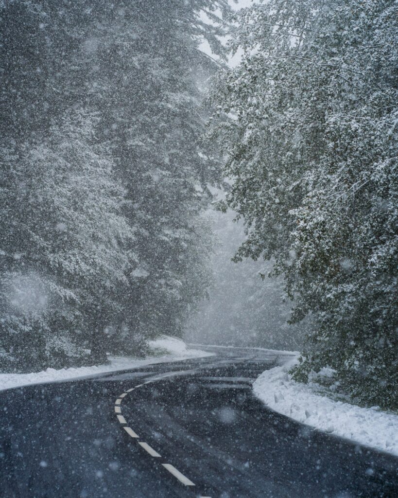 Winding snowy road through dense forest in heavy winter blizzard with white and gray tones Wallpaper