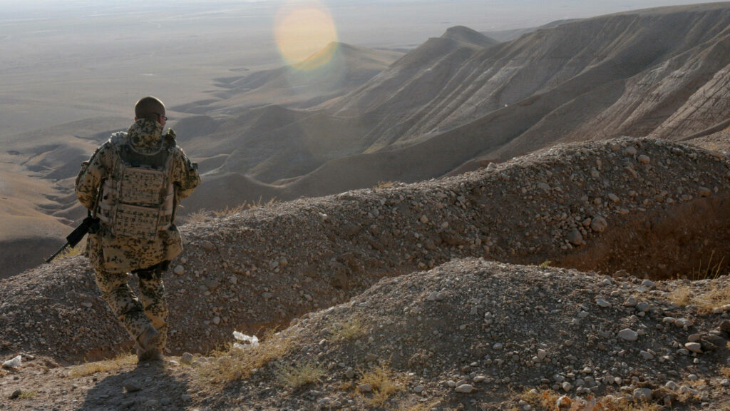 Soldier in camouflage gear on rugged Afghan mountain ridge overlooking vast arid landscape Wallpaper
