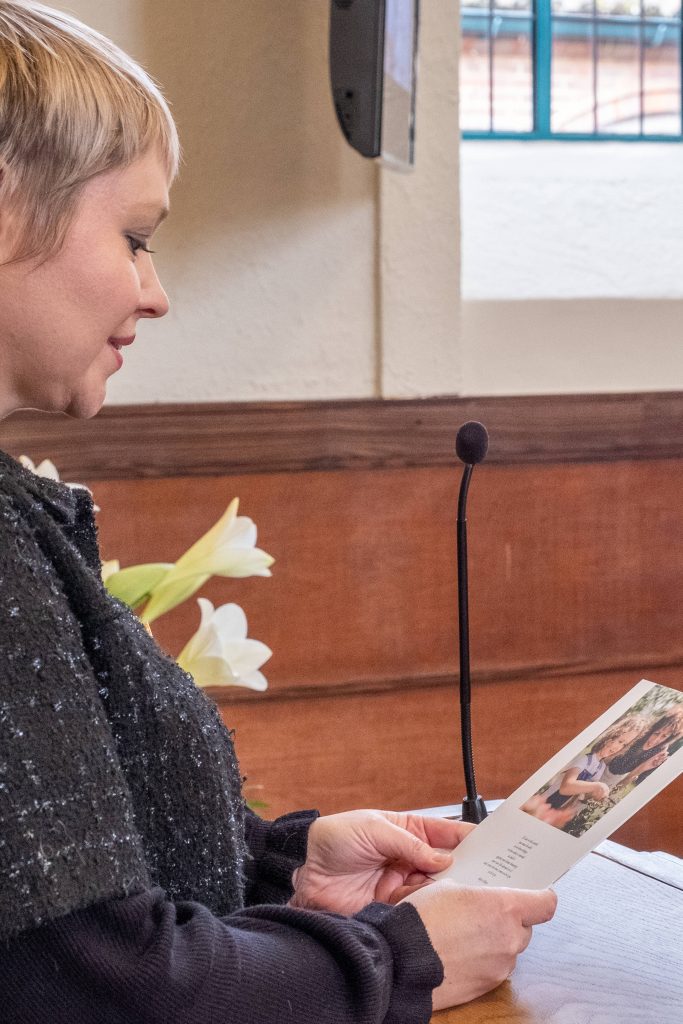 Woman in black sweater reading memorial program at podium with white lilies funeral wallpaper