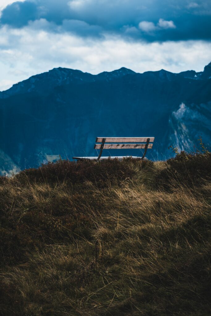 Solitary bench on grassy hill overlooking majestic mountain range under dramatic sky wallpaper