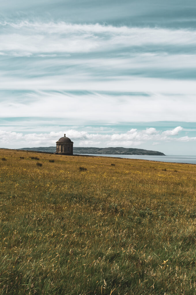 Solitary building on grassy field under dramatic clouds with distant hills and calm water horizon Wallpaper