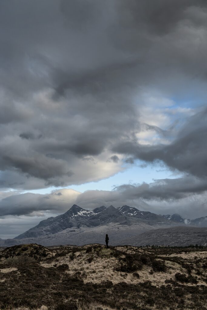 Lone figure under dramatic clouds with rugged mountain silhouette in vast natural landscape wallpaper