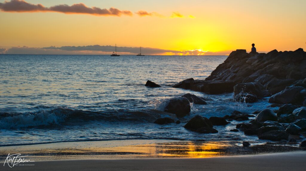 Solitary person sits on rugged rocks at sunset with waves shore sailboats serene sea blues golden hues Wallpaper