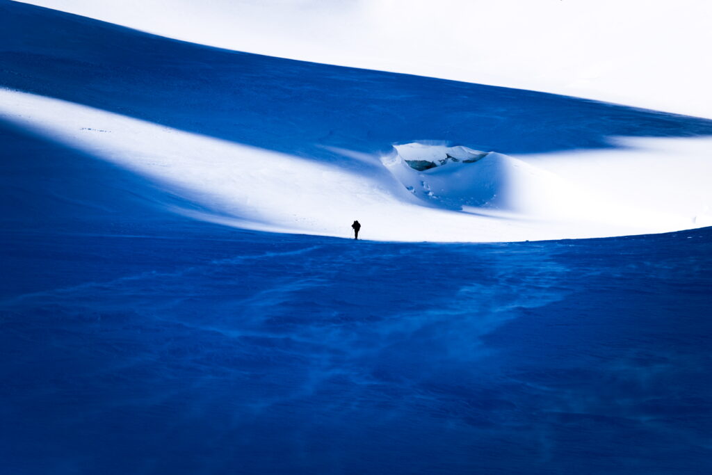 Solitary figure stands before snowy mountains with deep blue shadows and bright white snow background Wallpaper
