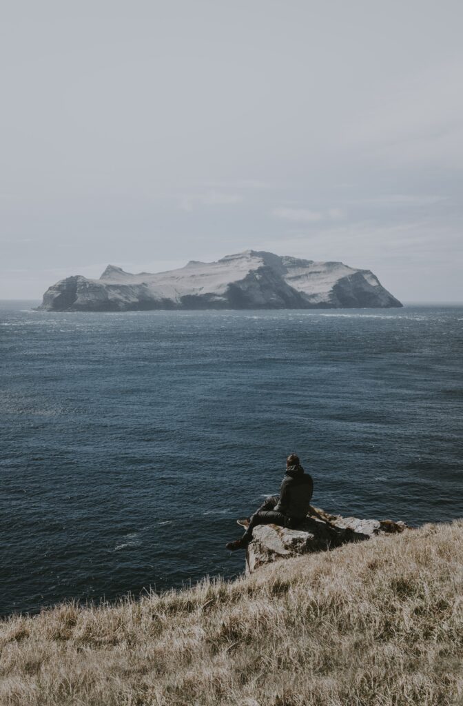 Solitary person on cliff edge overlooking calm sea and distant rugged island under overcast sky Wallpaper