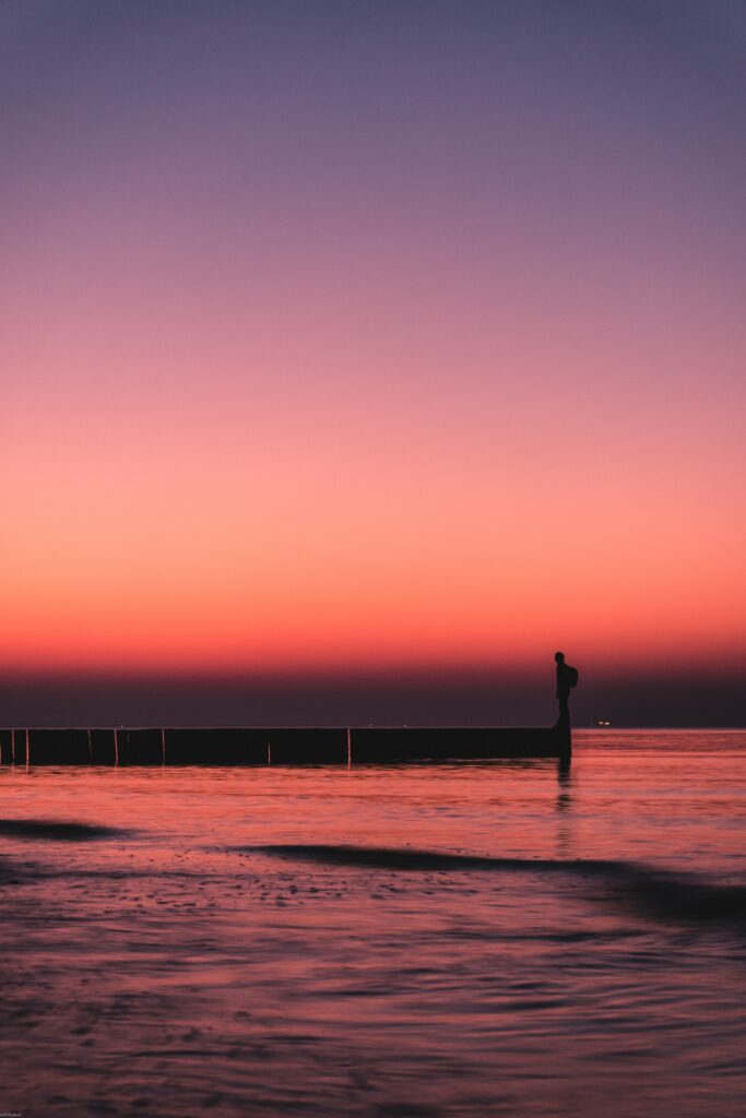 Solitary figure on pier gazing at vibrant sunset sea with purple orange sky and calm waters Wallpaper