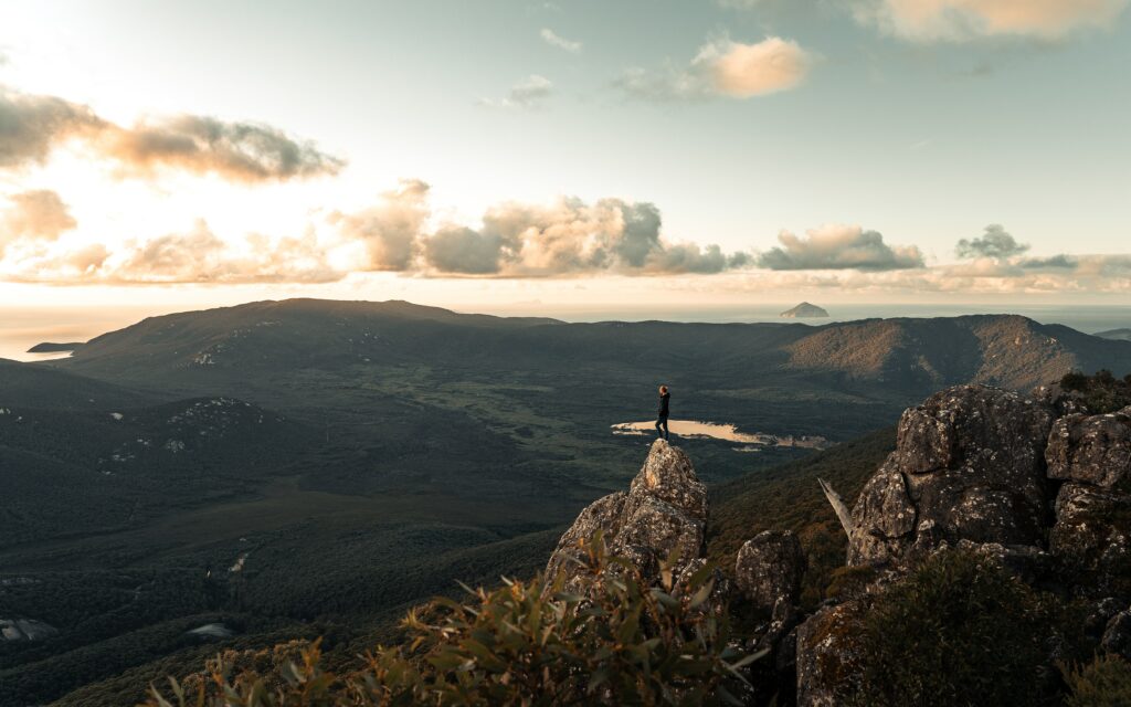 Lone figure on rocky outcrop overlooking green mountains and serene valley at golden sunset Wallpaper