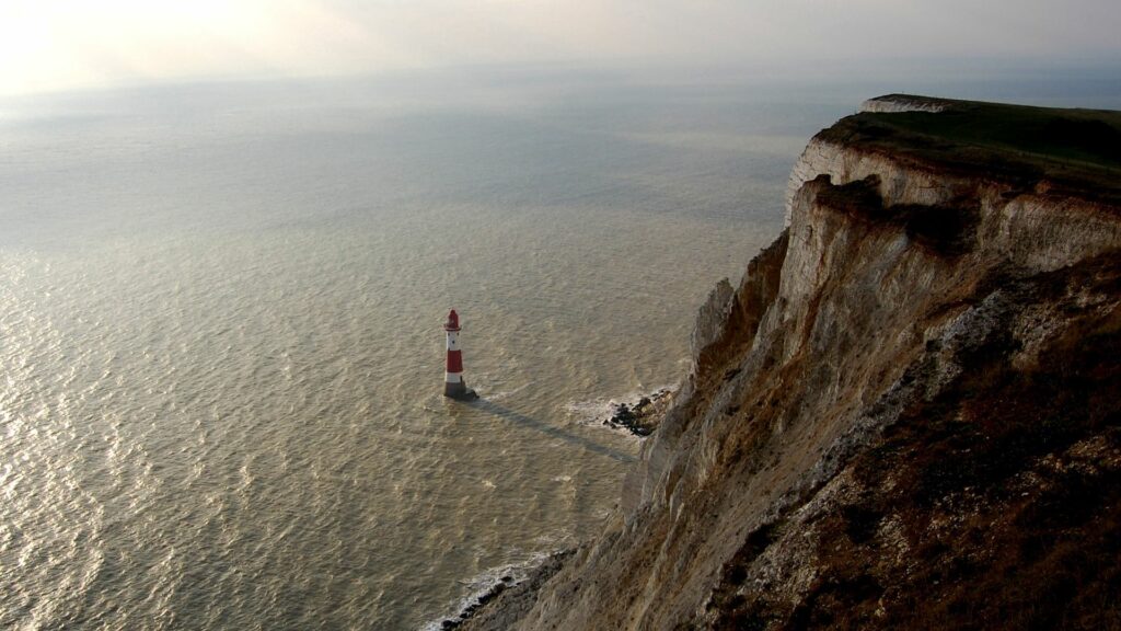 Solitary lighthouse by rugged cliff with waves crashing below under dramatic natural light wallpaper