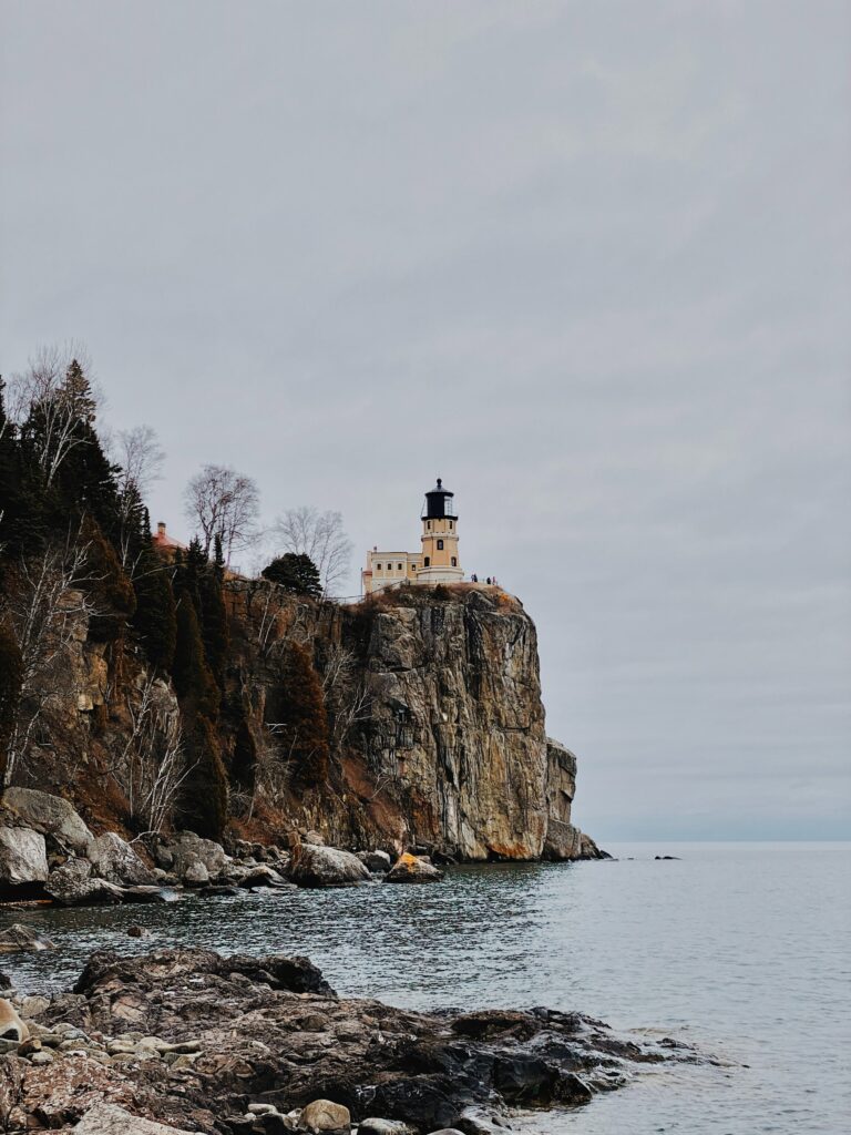 Solitary lighthouse on rugged cliff overlooking calm sea and rocky shore with muted earth tones Wallpaper
