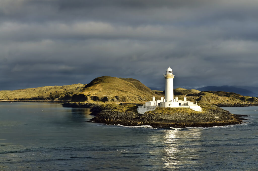 Solitary lighthouse on rugged island shore with calm blue sea golden hills and cloudy sky Wallpaper