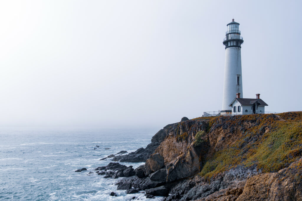 Solitary white lighthouse on rocky slope overlooking misty blue gray ocean coastal background Wallpaper