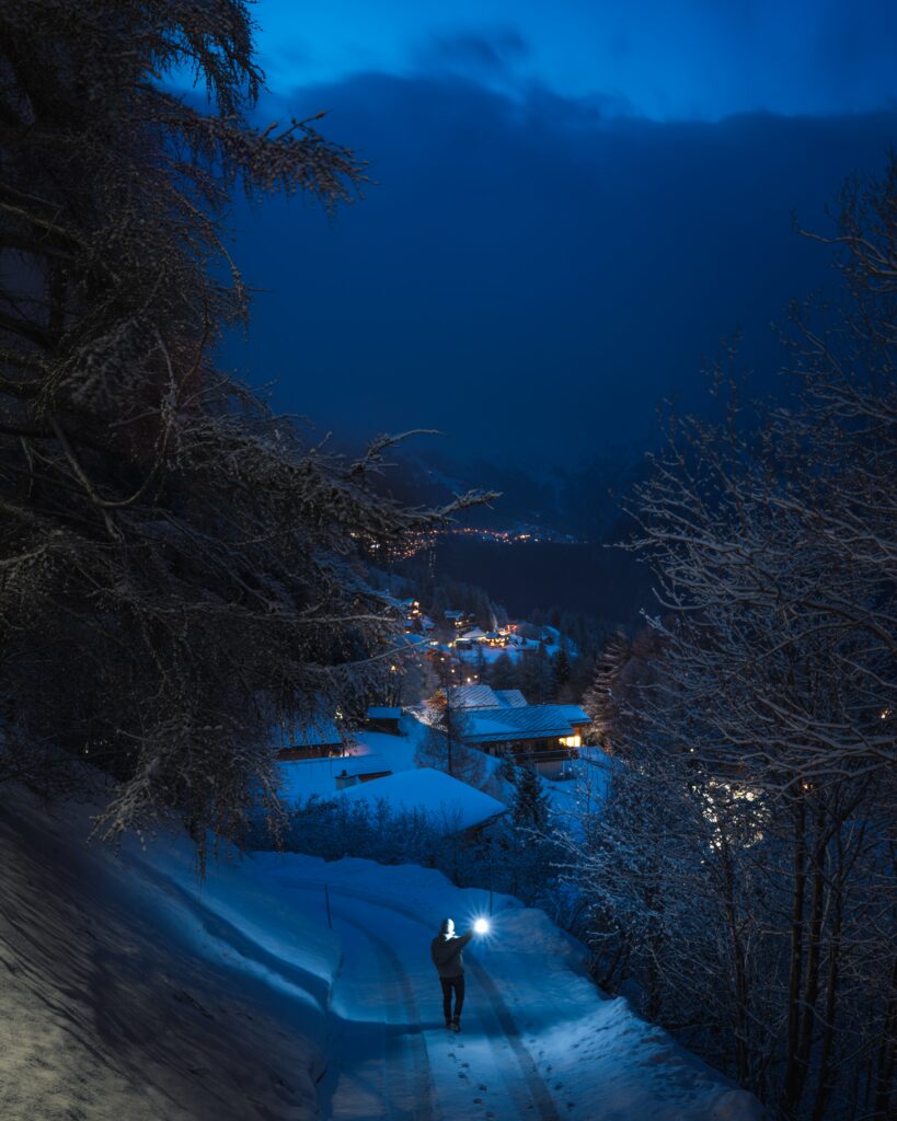 Solitary man walks snowy path winter evening frosted trees glowing village lights moody sky fog Wallpaper