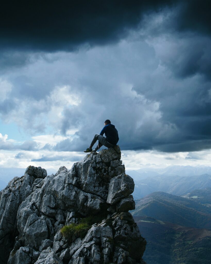 Man sitting on jagged mountain rock with dramatic clouds and vast sky creating solitude and freedom Wallpaper
