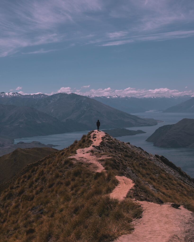 Man standing on rugged mountain ridge overlooking valleys and snow-capped peaks under serene sky Wallpaper