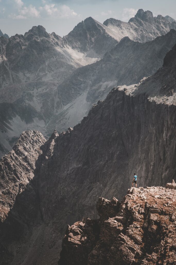 Man on rugged mountain rocks beneath towering peaks and hazy sky showcasing nature's majesty Wallpaper