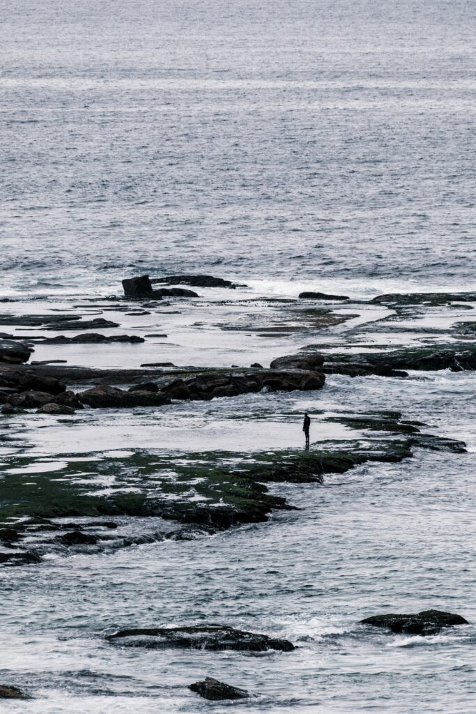 Solitary man on rugged rocks with crashing ocean waves moody gray blue seascape wallpaper background