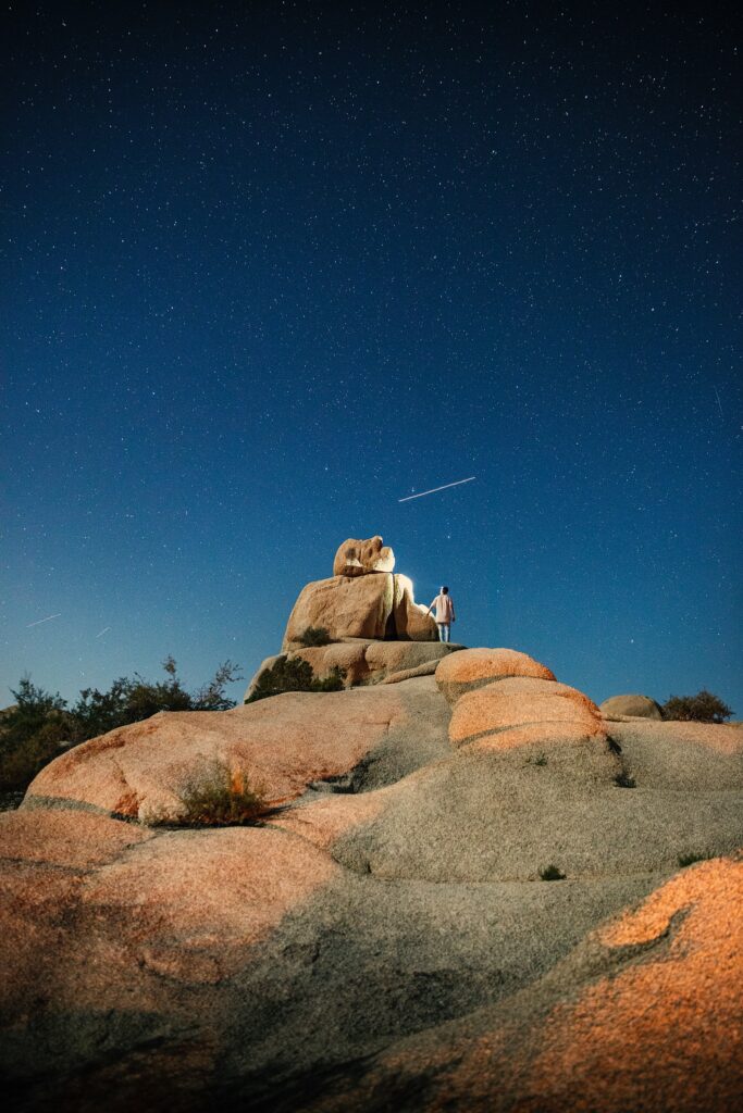 Man standing on rugged rocks under starry night sky serene nature solitude wallpaper background