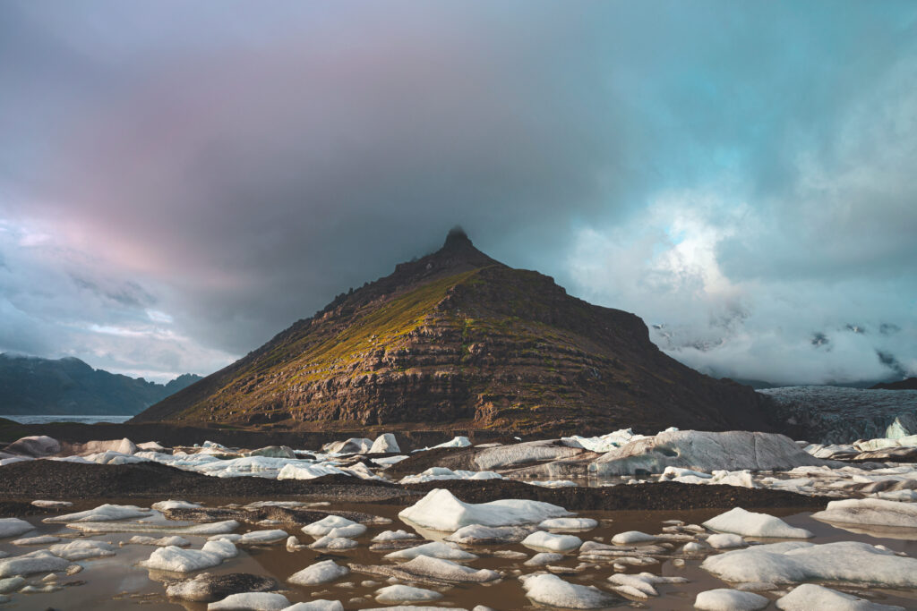 Rugged green hill with snow patches beneath dramatic clouds in serene natural landscape wallpaper