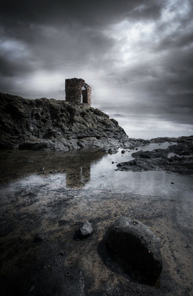 Solitary stone ruin on rocky shore with moody clouds and reflective still water wallpaper background
