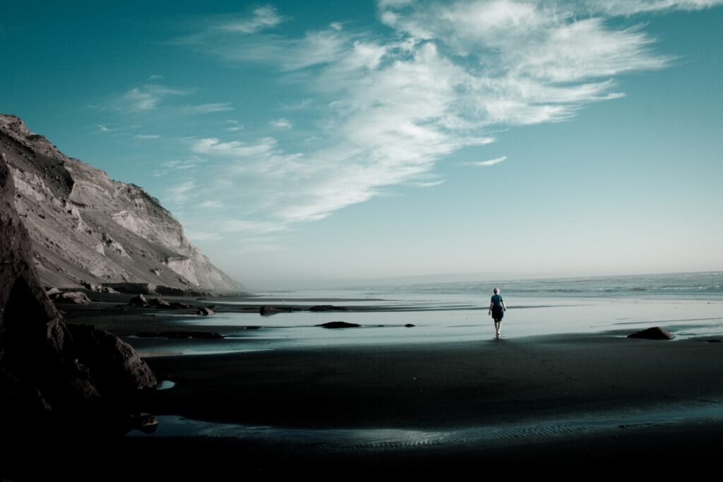 Solitary figure walks on dark sandy beach with cliffs under dramatic cloudy sky wallpaper background