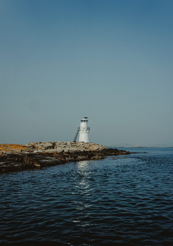 White lighthouse on rocky shore overlooking calm blue sea under clear sky minimalist coastal scene Wallpaper