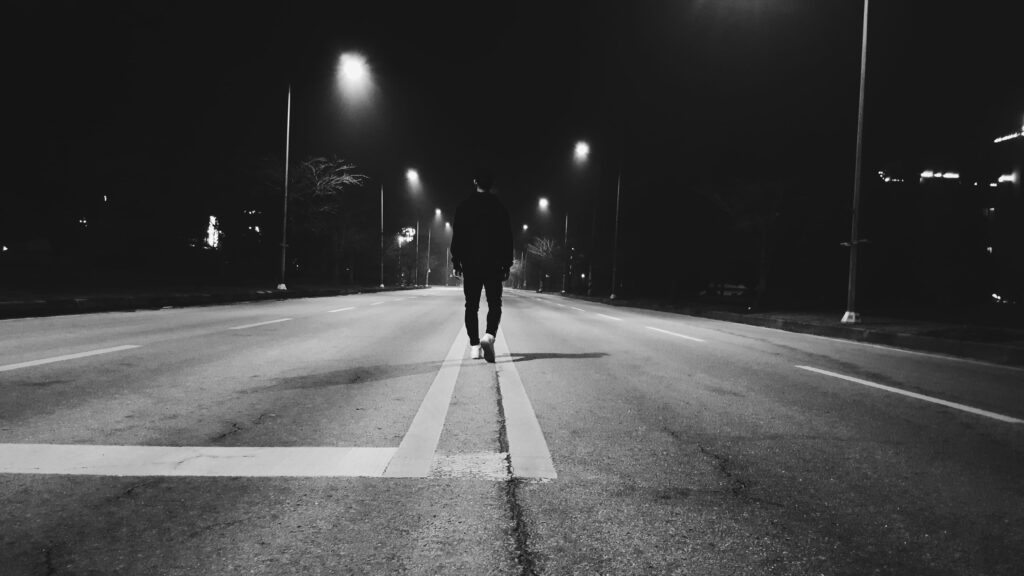 Solitary Stroll: A Man Walking Alone in the Black Night, Illuminated by the Light of Transportation on the Road - Wallpaper Background Photo