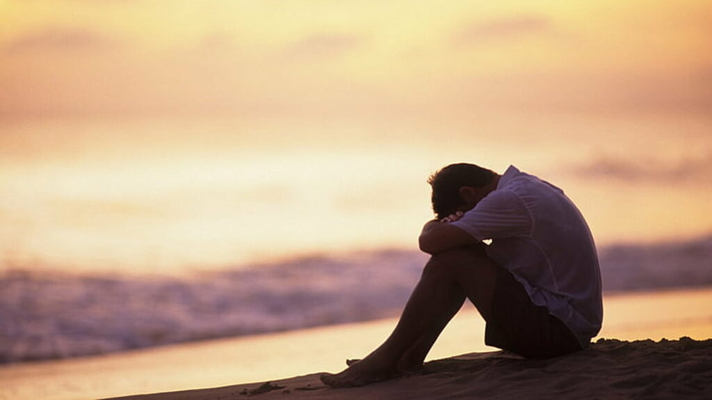 Man sitting alone on beach at sunset with golden light evoking solitude and deep reflection wallpaper