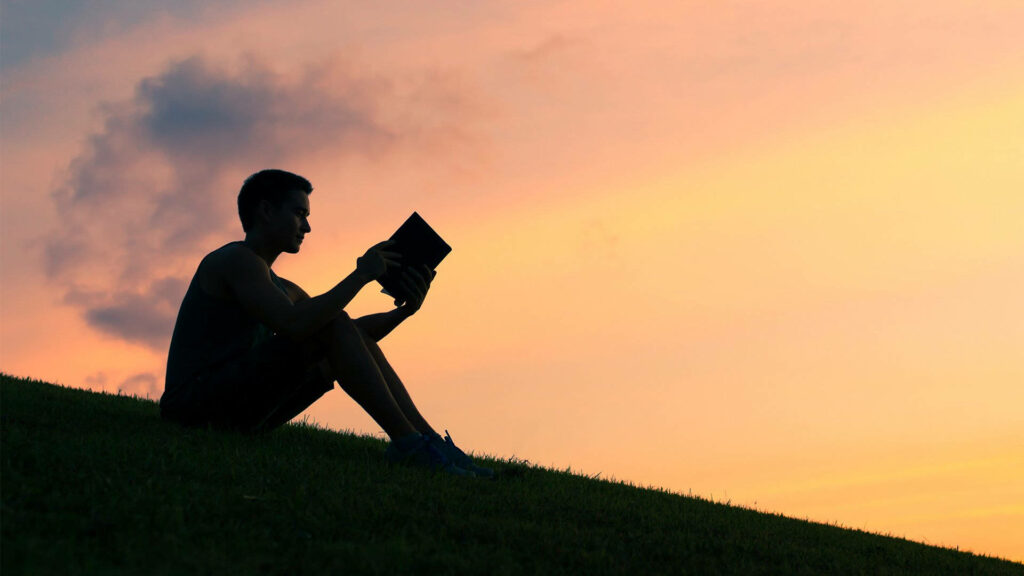 Solitude in the Scenic Splendor: Captivating Background Portrait of a Solitary Young Man Immersed in a Book Amidst Hilltop Serenity at Sunset Wallpaper